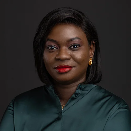 Evelyne Dioh Simpa, portrait en buste sur fond gris foncé, chemisier vert émeraude et boucles d’oreilles dorées